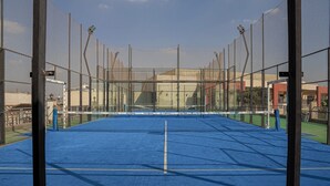 Una cancha de tenis con red, rodeada por una valla, y un edificio al fondo.