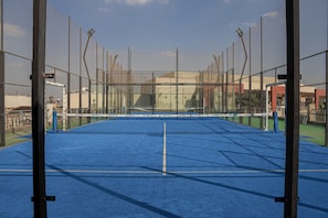 A tennis court with a net, surrounded by a fence, and a building in the background.