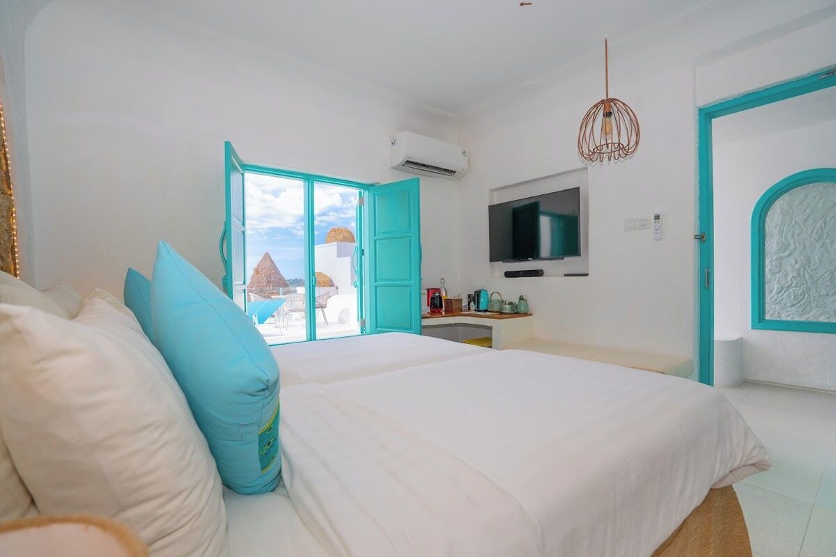 Ein Schlafzimmer mit Bett, Fernseher, Schreibtisch und Blick auf ein Gebäude mit einer pyramidenförmigen Dachkonstruktion.