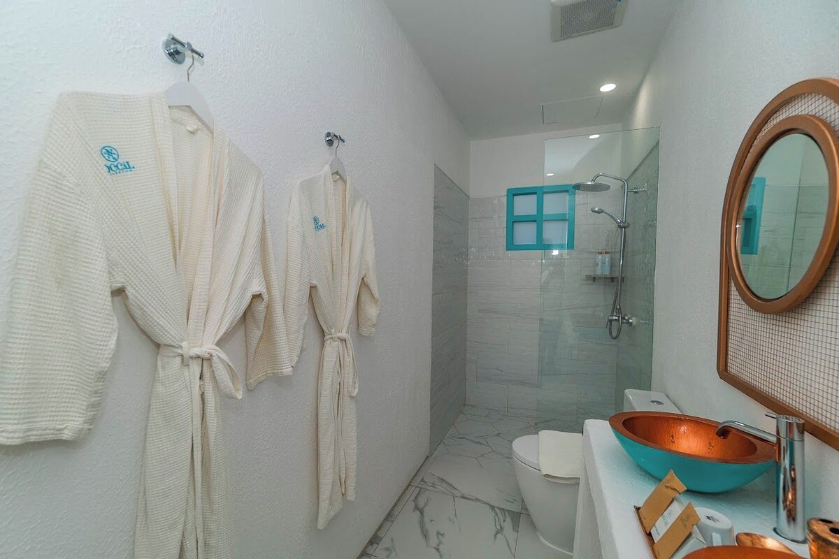 Ein modernes Badezimmer mit Duschkabine aus Glas, einem weißen Waschbecken mit Kupferbecken und drei weißen Bademänteln, die an der Wand hängen.