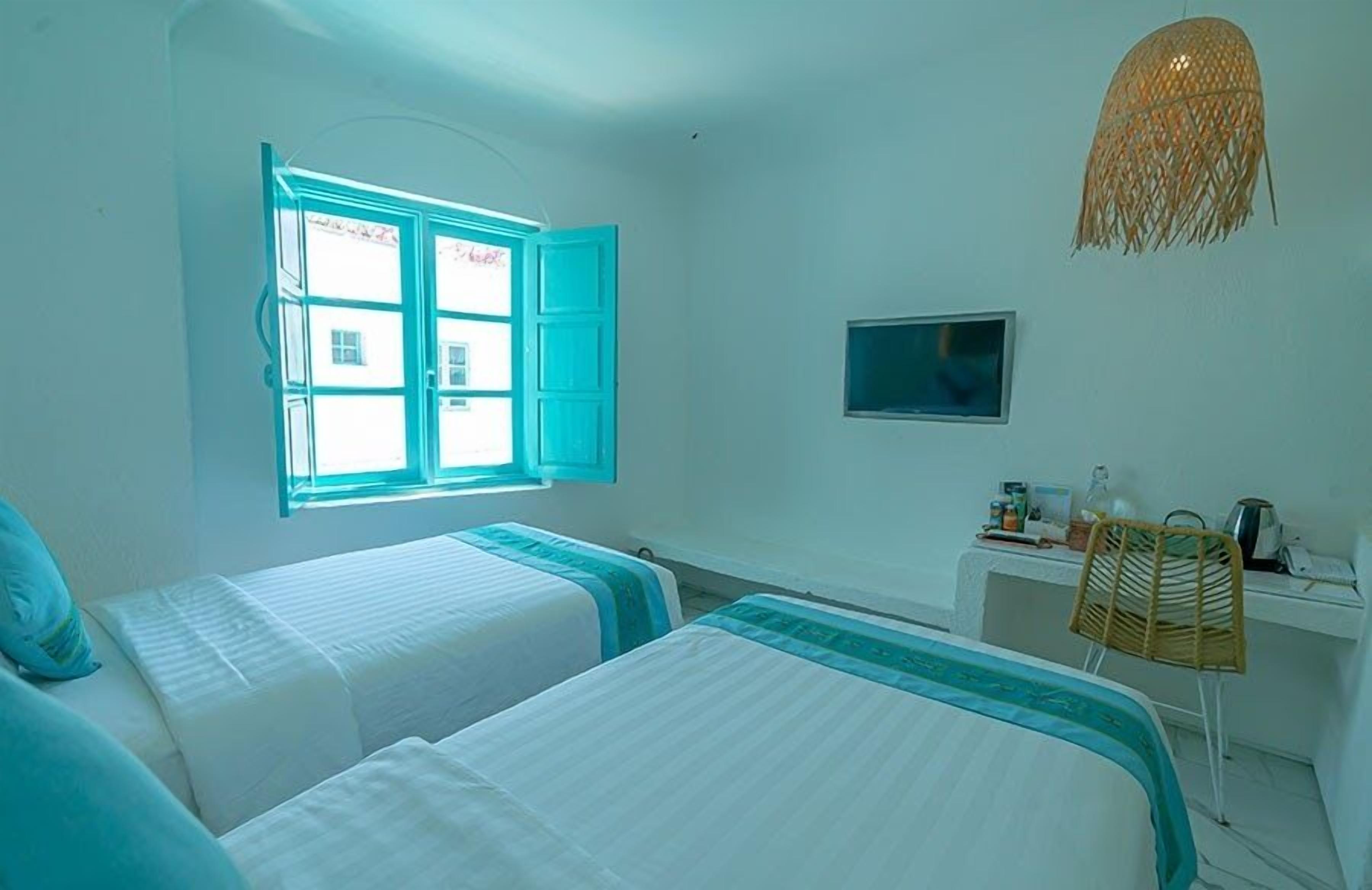 A room with two beds, a desk, a chair, a television, and a window with open shutters.