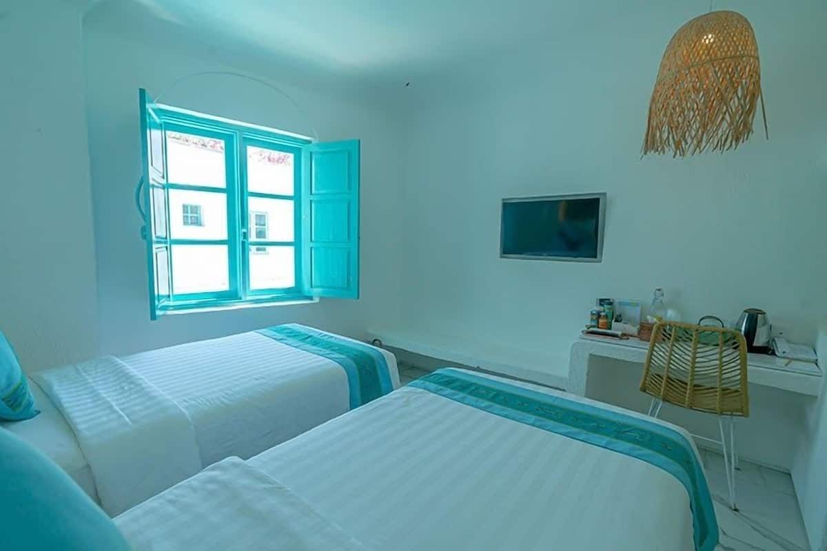 A room with two beds, a desk, a chair, a television, and a window with open shutters.