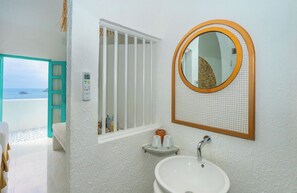 A bathroom with a white sink, a round mirror with a wooden frame, a window with white bars, and a view of the sea. - Loccal Collection Hotel Labuan Bajo (Labuan Bajo)