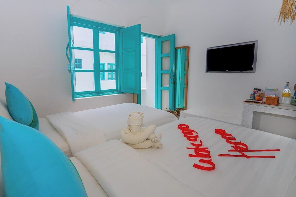 A hotel room with a bed, a TV, a window with an open shutter, and a small table with items on it.