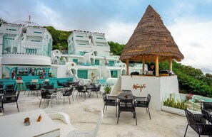 Un bar en la azotea con mesas al aire libre, una estructura con techo de paja y edificios blancos con detalles azules.