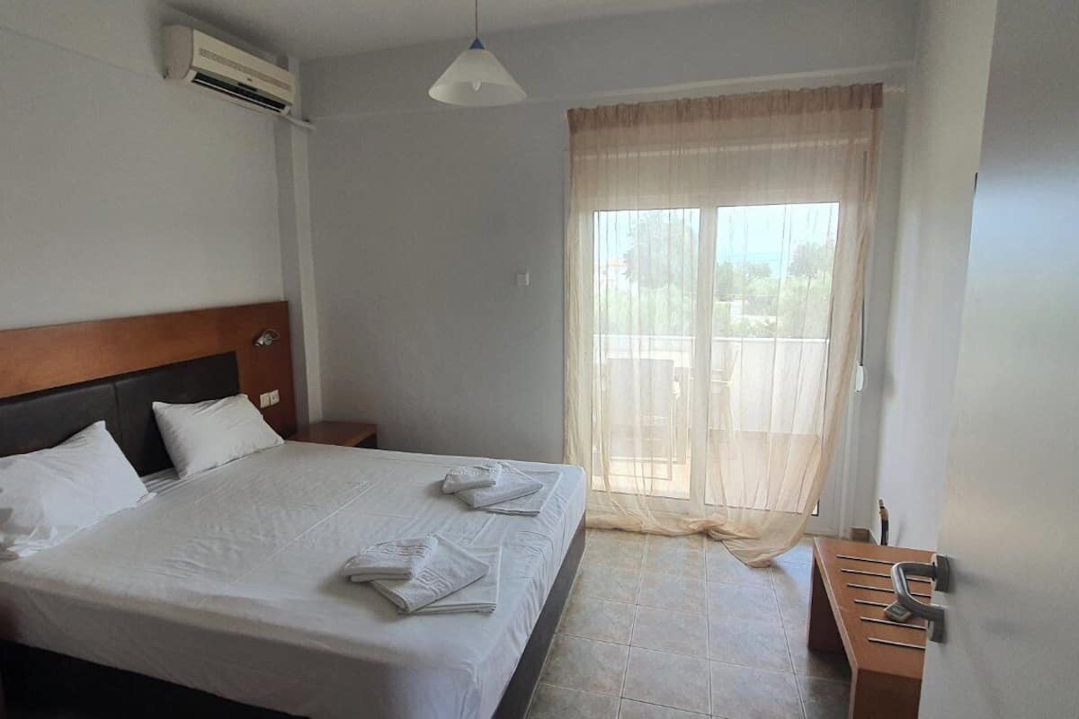 A hotel room with a bed, a nightstand, a wooden headboard, a window with sheer curtains, and a view of the outdoors.