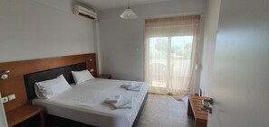 A hotel room with a bed, a nightstand, a wooden headboard, a window with sheer curtains, and a view of the outdoors. - Villa Ilios Hotel (Kassandra)