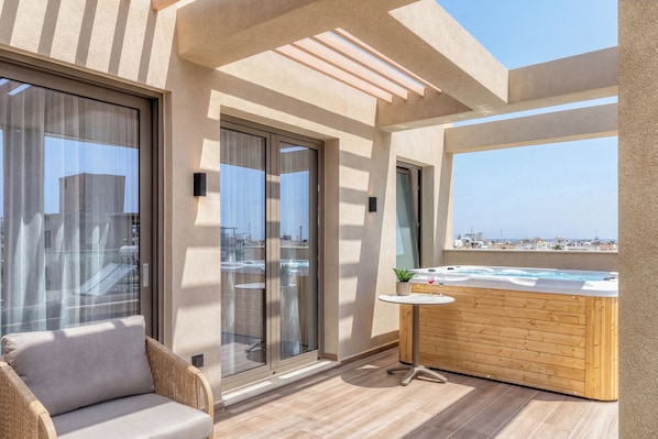 A balcony with a wooden hot tub, a cushioned chair, and a view of the city.