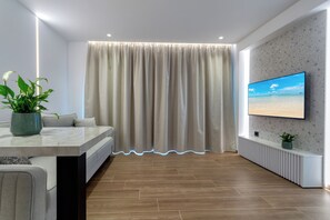 A modern living room with a sofa, a dining table, a TV mounted on the wall, and a large window with curtains. - Lemar (Hersonissos)