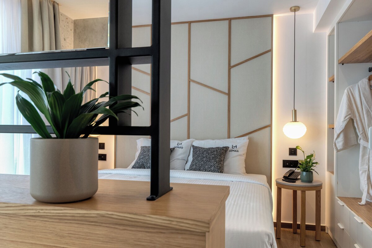 Une chambre d’hôtel moderne, équipée d’un lit, d’une table de chevet avec une plante, d’une petite table d’appoint avec une lampe et d’une armoire où sont suspendus des vêtements.