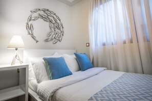A neatly made bed with blue and white bedding, a white nightstand with a lamp, and a wall art piece above the bed. - Lemar (Hersonissos)