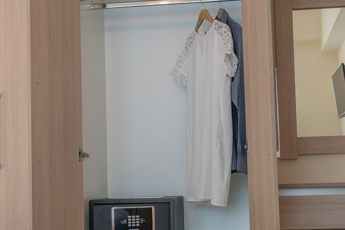 A hotel room with a bed, a wardrobe with hanging clothes, a desk with a laptop, and a bedside table with a small appliance.