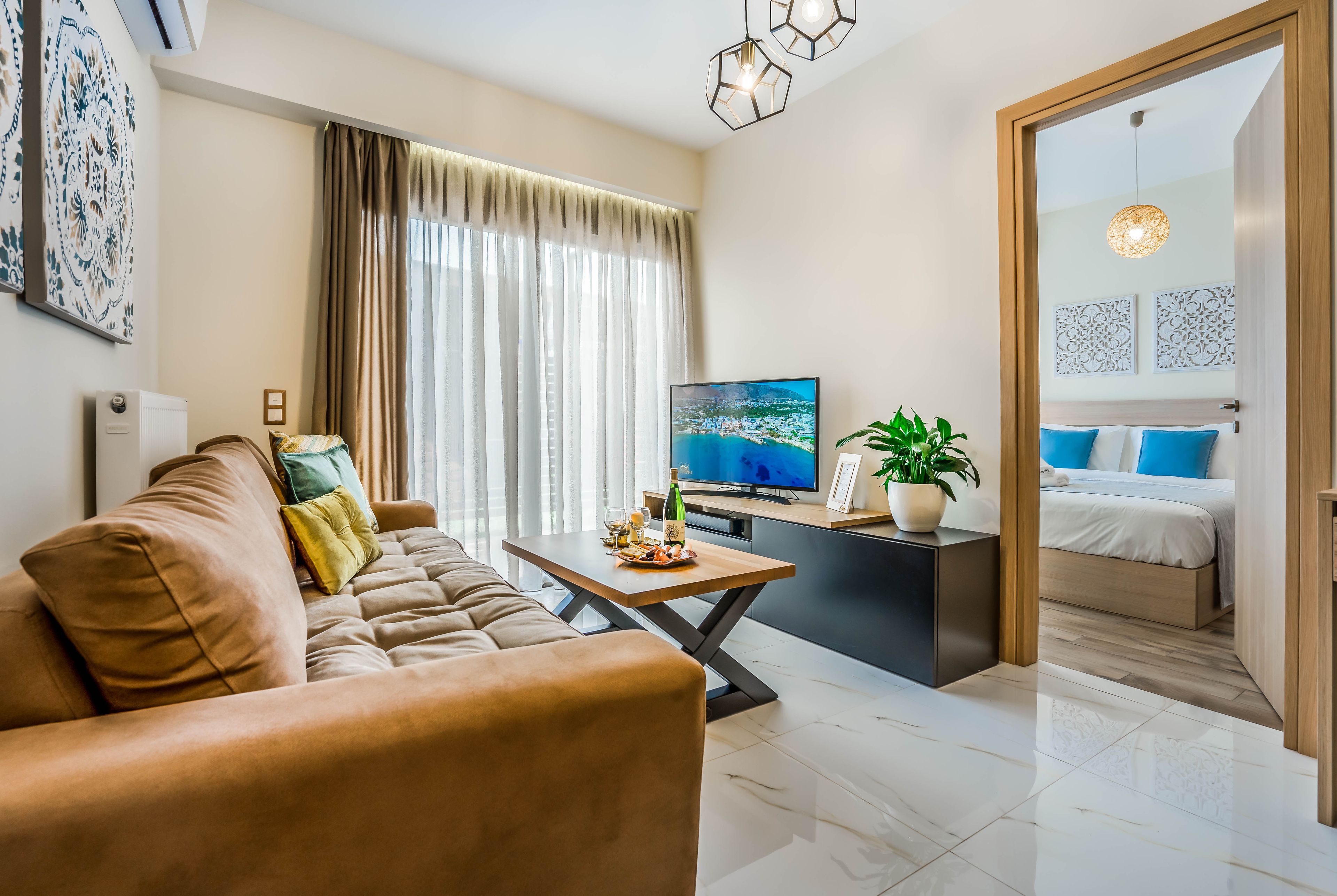A modern living room with a sofa, a coffee table with a bottle and glasses, a TV, and a bed with blue pillows.