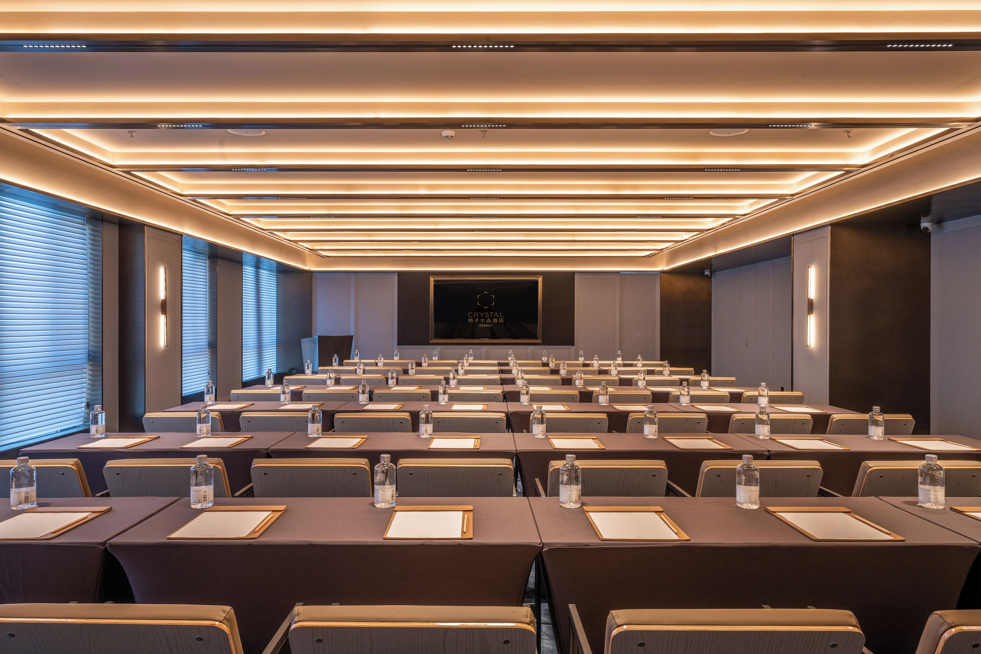 A conference room with rows of tables, chairs, and water bottles, featuring a modern ceiling design and a central screen.