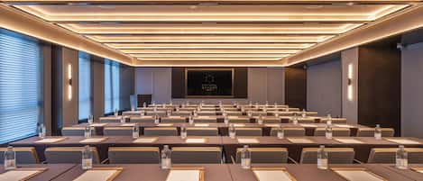 Un salón de conferencias con filas de mesas, sillas y botellas de agua, que presenta un diseño moderno en el techo y una pantalla central.