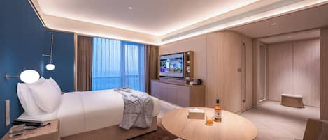 Quarto de hotel moderno com uma cama grande, televisão de tela plana, mesa de centro redonda e área de estar com sofá e poltronas.