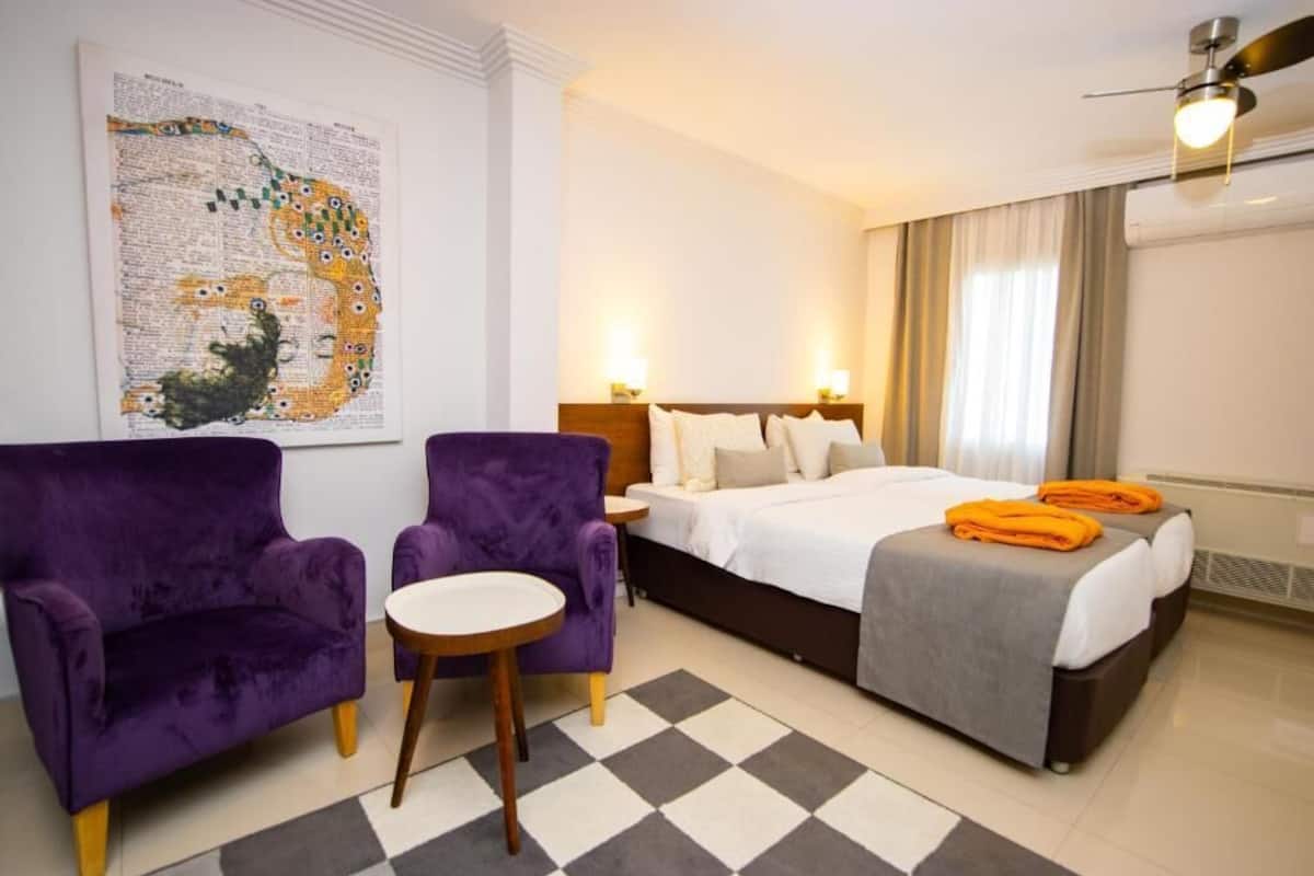 Ein Hotelzimmer mit einem Bett, zwei violetten Sesseln, einem kleinen runden Tisch, einem Schachbrettteppich und einem Wanddekor.