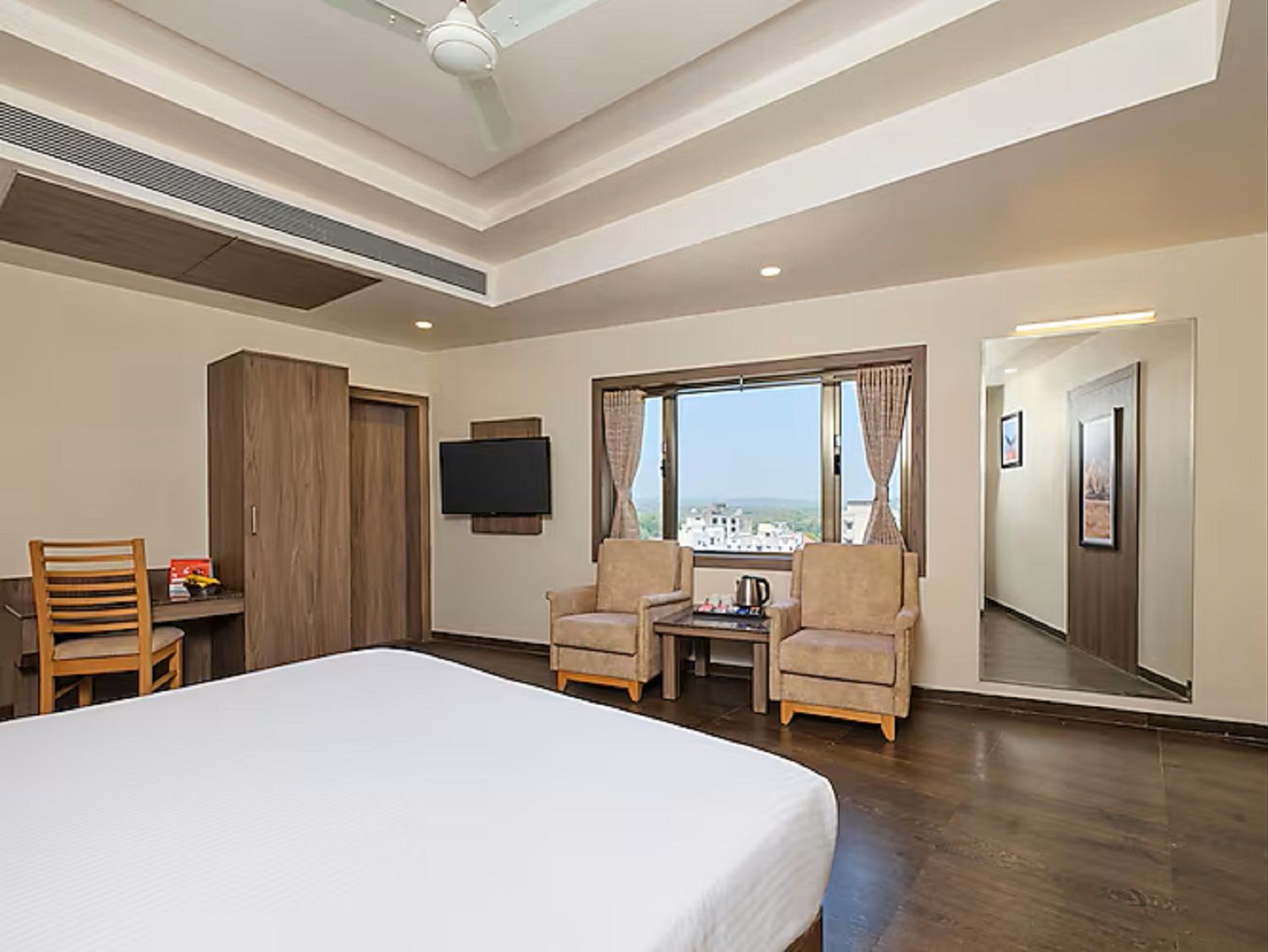 A hotel room with a bed, two armchairs, a small table, a TV, and a large window offering a city view.