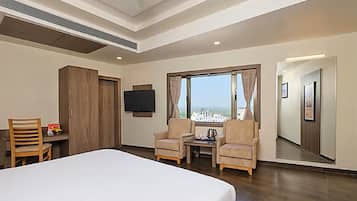 A hotel room with a bed, two armchairs, a small table, a TV, and a large window offering a city view.