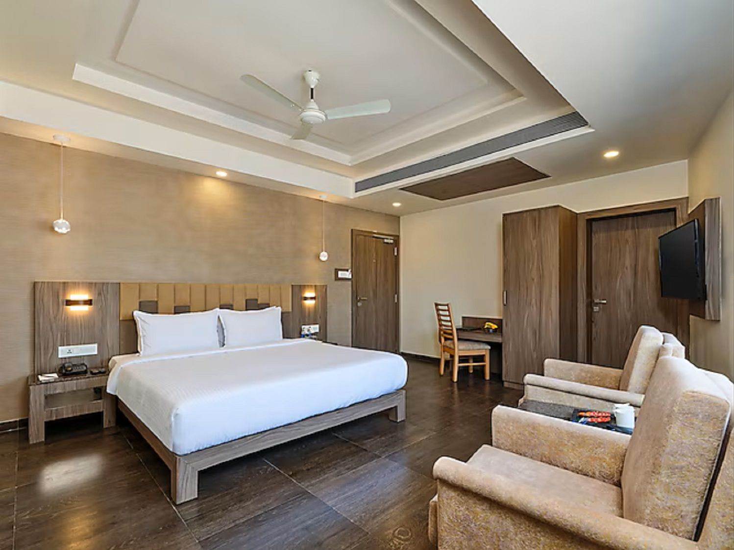 Quarto de hotel com uma cama grande, escrivaninha, cadeira, televisão e ventoinha no teto.