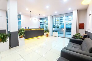 Vestíbulo moderno de hotel con mostrador de recepción, zona de estar y grandes ventanales que ofrecen vistas del exterior del edificio.