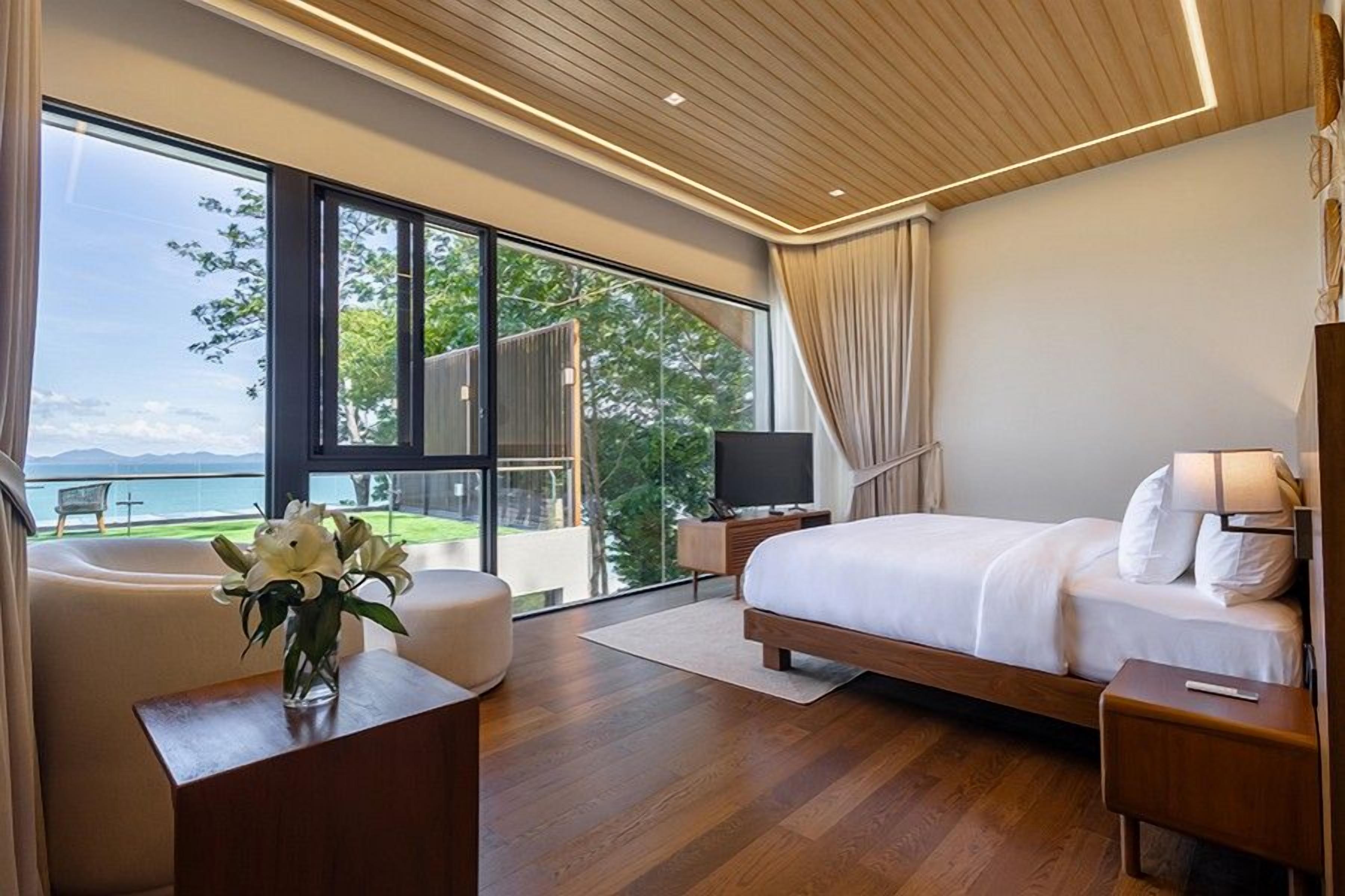 A modern bedroom with a large bed, a TV, a desk, and a view of the outdoors.