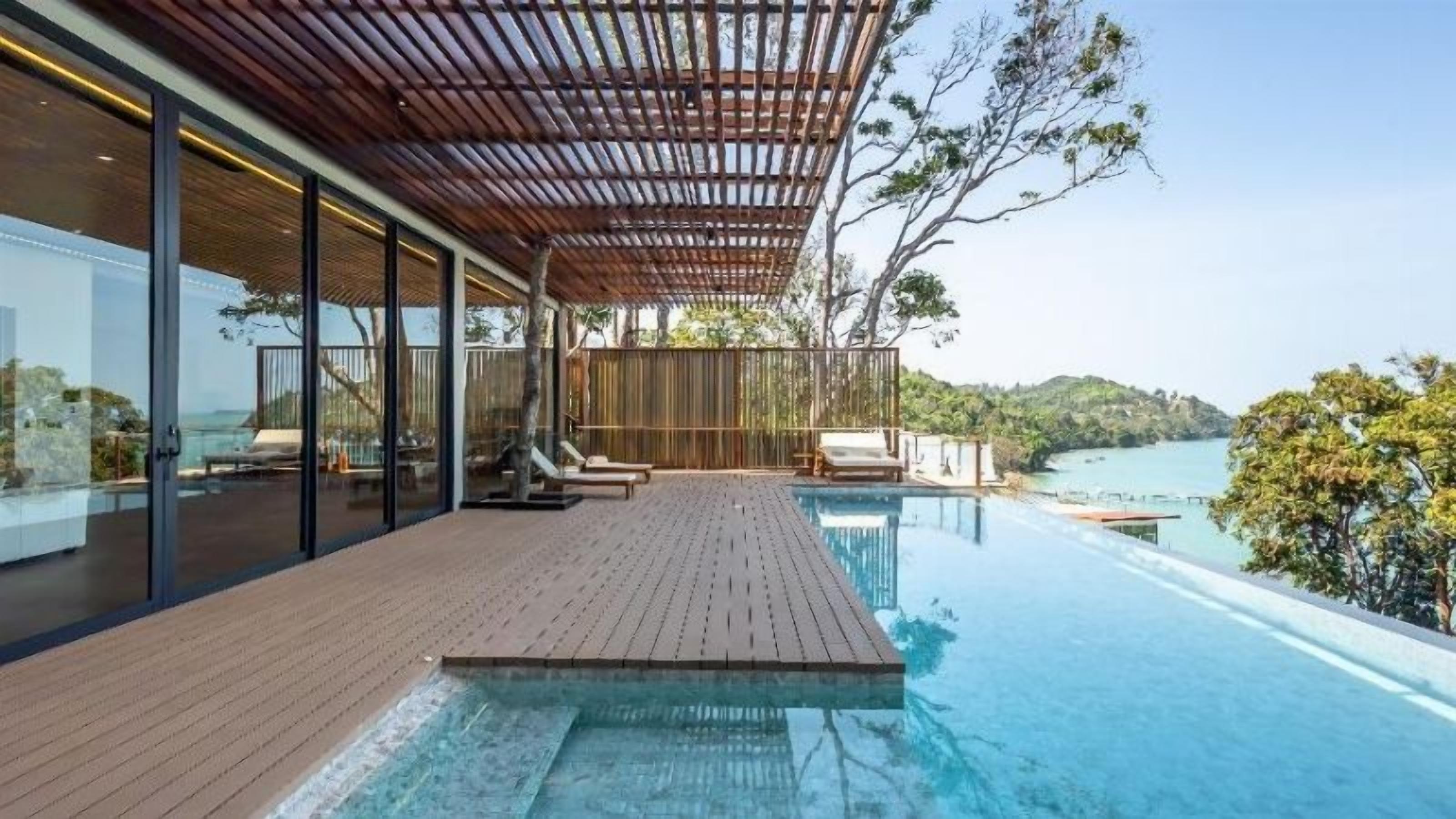 A modern infinity pool with a wooden deck, glass walls, and a view of the sea.