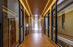 Um corredor com portas e janelas de vidro, piso de madeira e teto com iluminação linear.