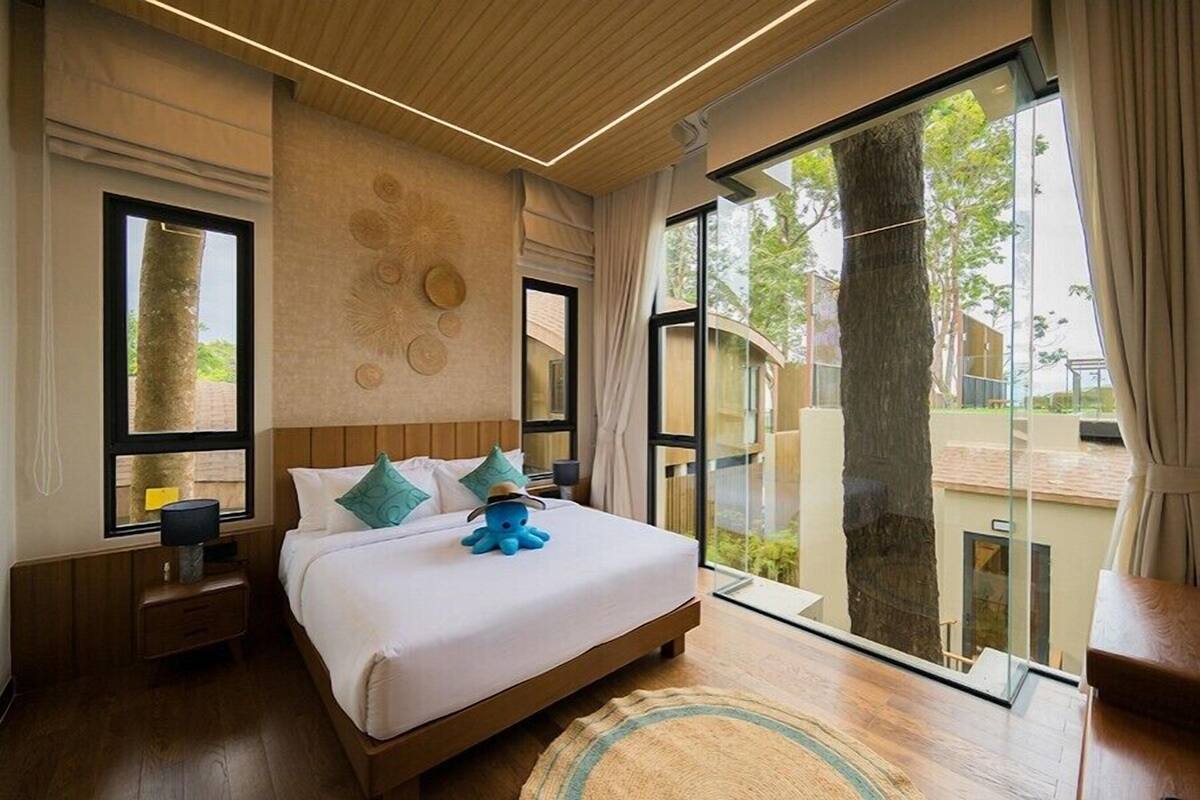 Habitación de hotel moderna con una cama grande, una mesita de noche, una lámpara y vistas al exterior a través de amplias ventanas.