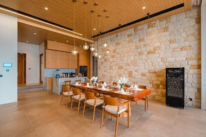 A modern dining area with a long wooden table, cushioned chairs, hanging spherical light fixtures, and a stone wall backdrop. - Sinae Residence Pool Villa (Ratsada)