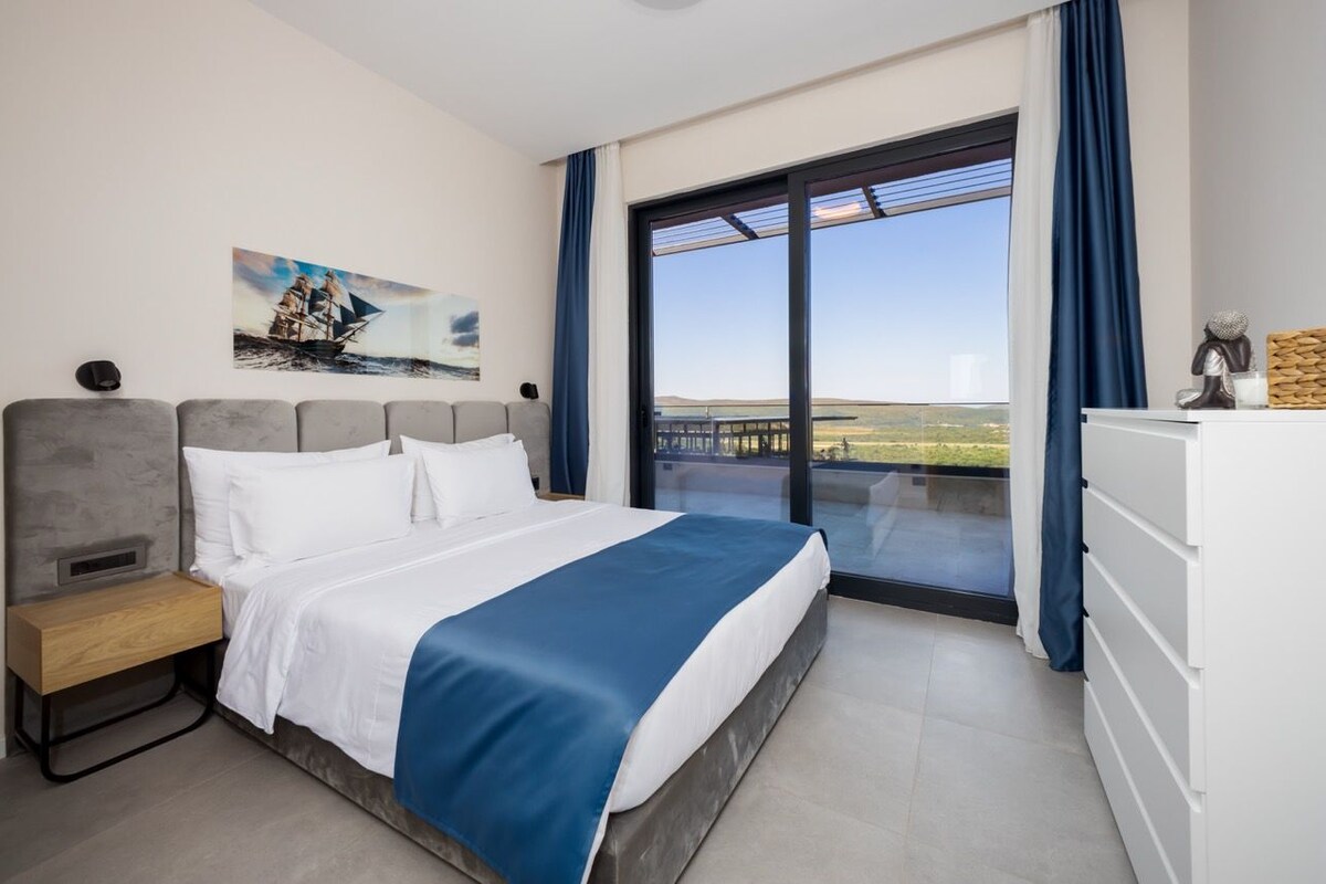 Une chambre à coucher comprenant un lit, une table de chevet, une commode, un tableau et une grande fenêtre donnant sur un beau paysage.