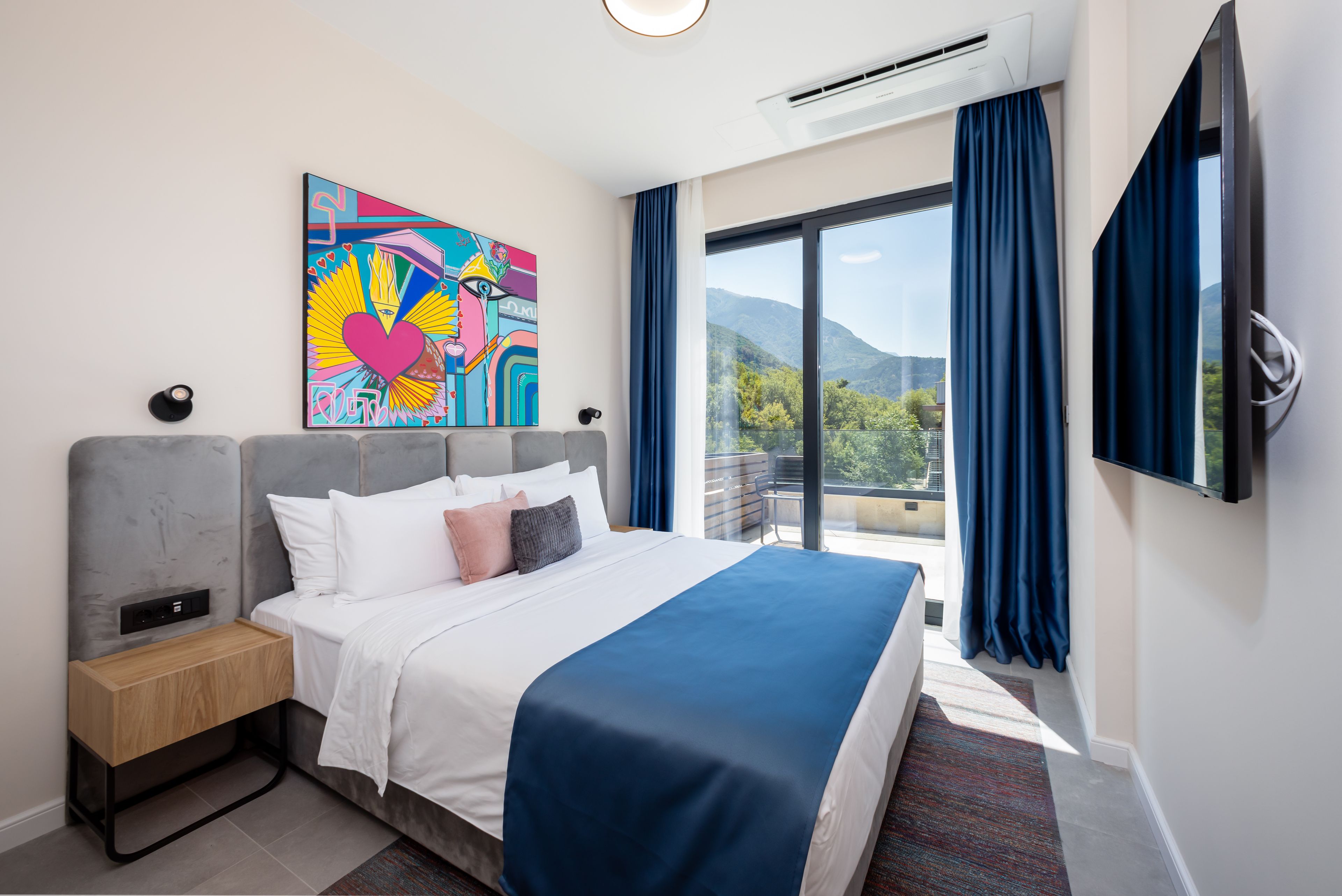 A hotel room with a bed, a nightstand, a painting, a TV, and a large window with curtains.