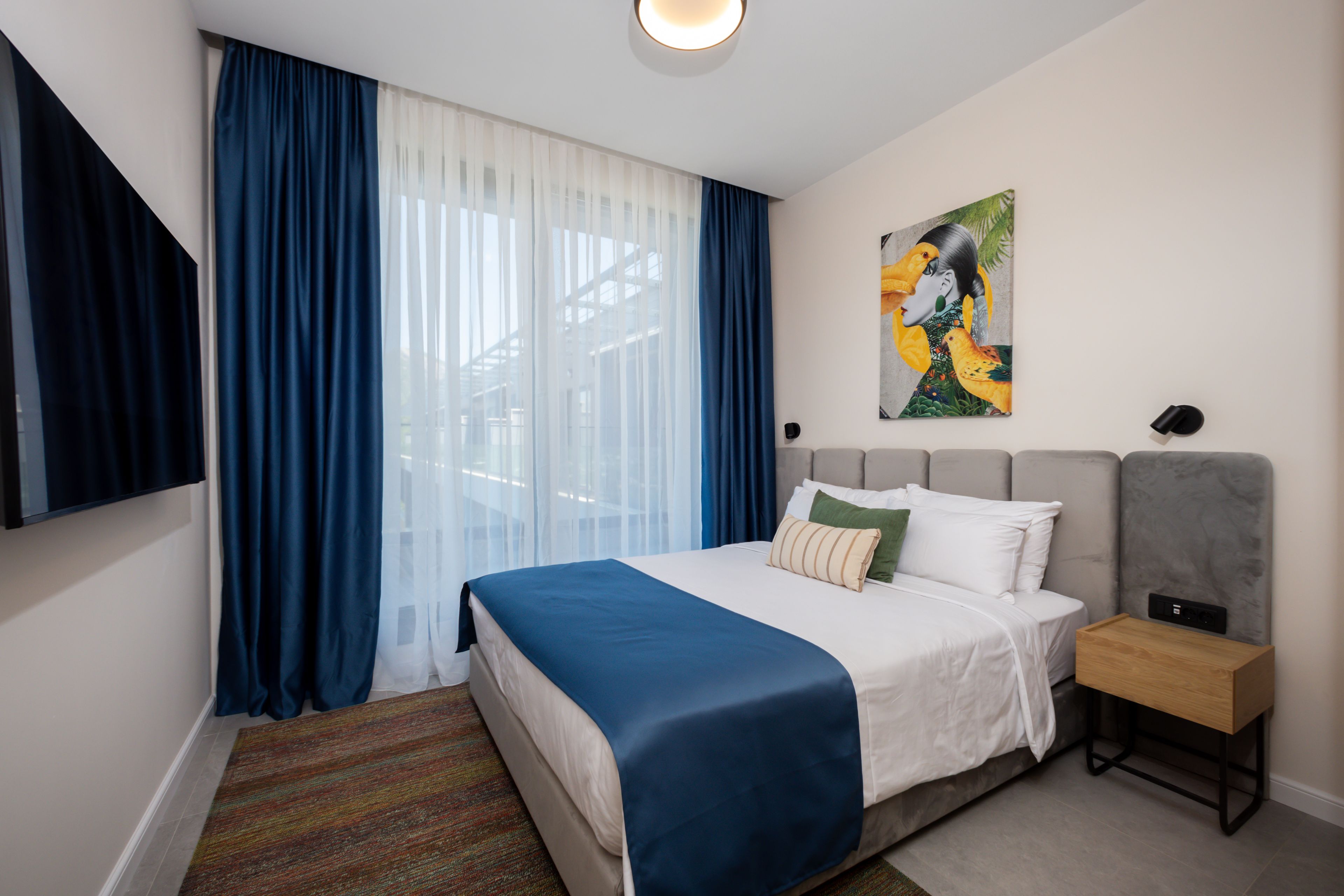 A hotel room with a bed, a nightstand, a painting, and a window with curtains.