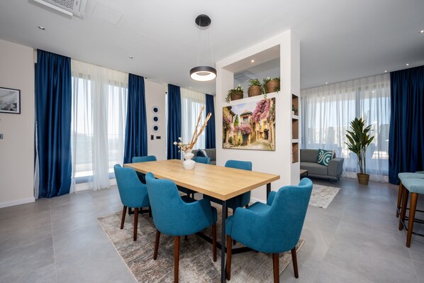 Una sala de estar moderna con comedor, que incluye una mesa de madera, sillas azules, un sofá y un amplio ventanal con cortinas.