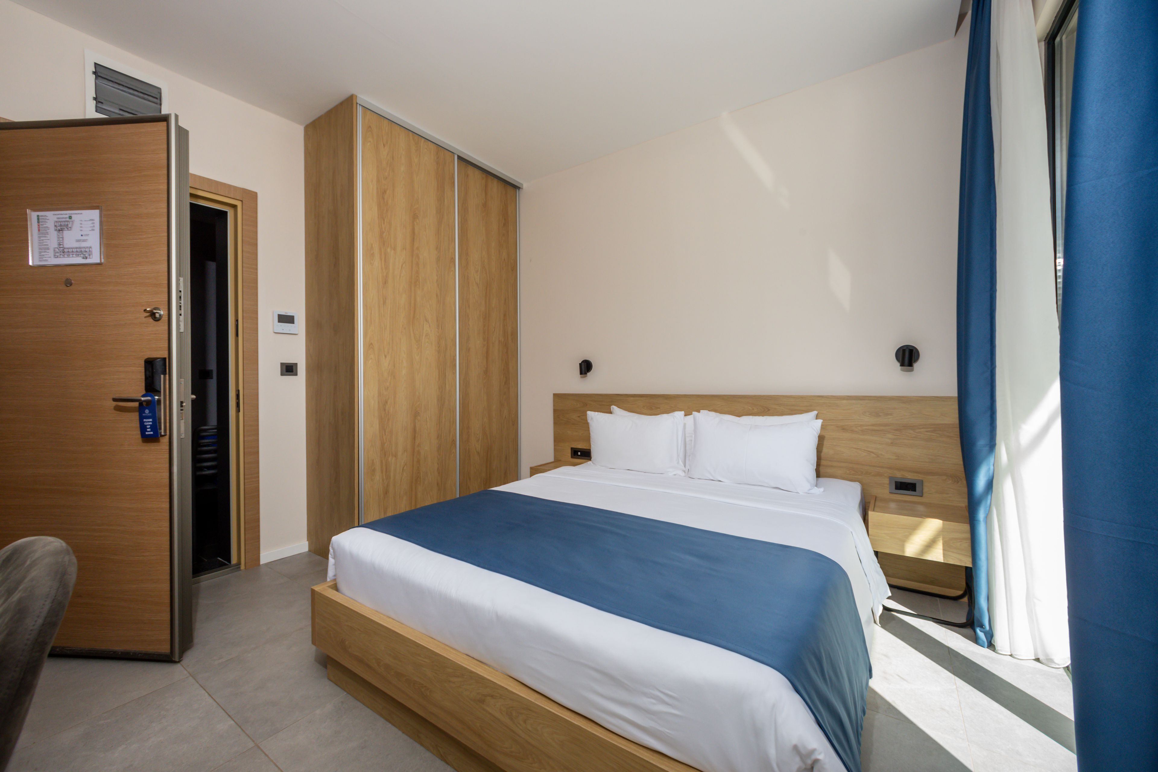 Quarto de hotel com cama, guarda-roupa, escrivaninha e janela com cortinas.