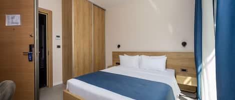 Una habitación de hotel con cama, armario, escritorio y ventana con cortinas.