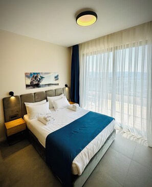 A hotel room with a large bed, a nightstand, a painting on the wall, and a window with curtains. - Royal Blue Resort And Residences (Kotor)