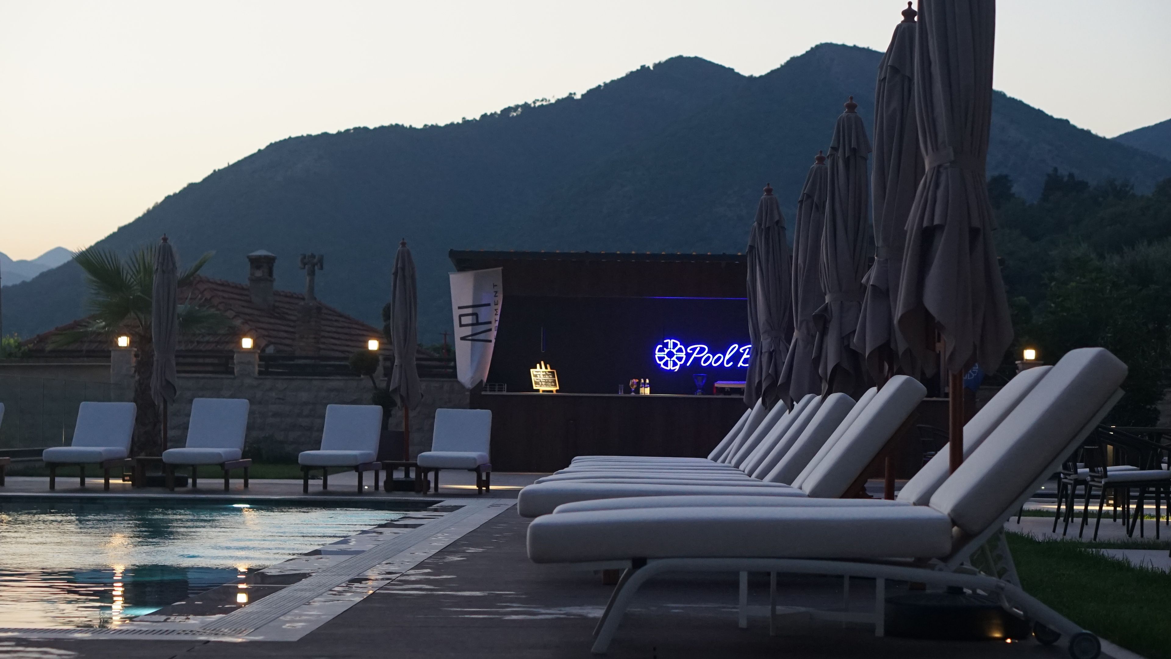 Zona junto a la piscina con sillones de descanso y sombrillas, un bar con letrero de neón y montañas al fondo.