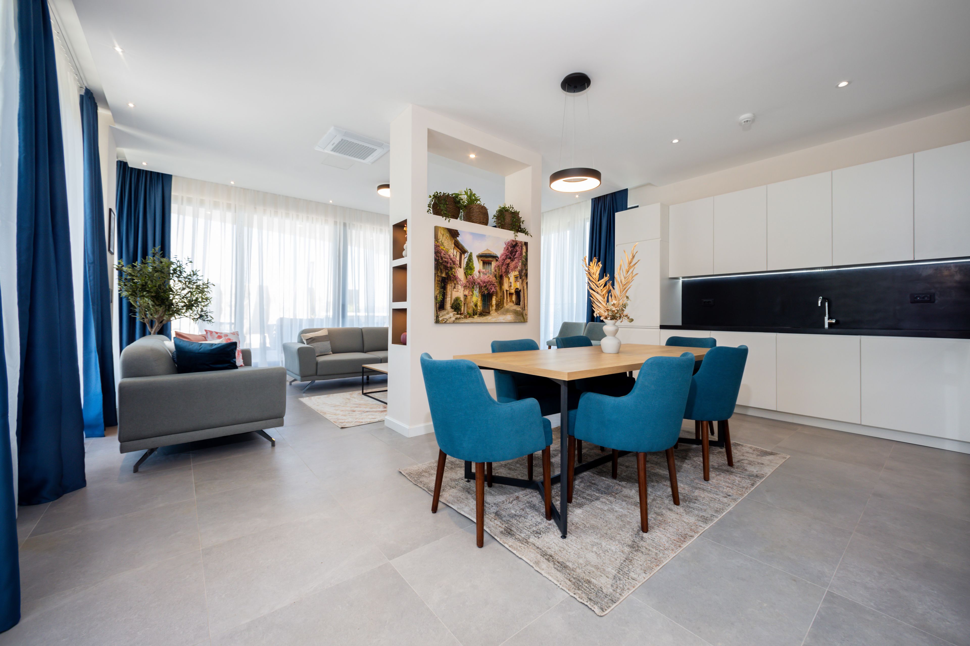 A modern living room with a sofa, a dining table with chairs, a kitchen area with cabinets, and a large window with curtains.