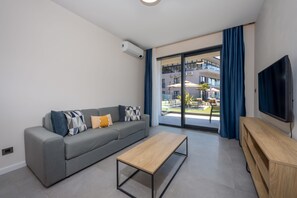 A modern living room with a grey sofa, a wooden coffee table, a TV, and a sliding glass door leading to an outdoor area. - Royal Blue Resort And Residences (Kotor)
