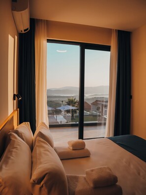 A hotel room with a bed, a window with curtains, a view of the landscape, and an air conditioning unit. - Royal Blue Resort And Residences (Kotor)