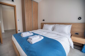 A neatly made bed with white linens and a blue accent, a wooden nightstand with a charging station, and a wardrobe with sliding doors. - Royal Blue Resort And Residences (Kotor)