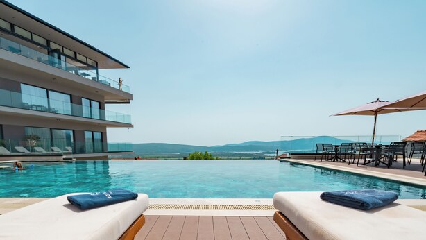 Un área de piscina con dos sillas reclinables, una terraza de madera y vistas a montañas.