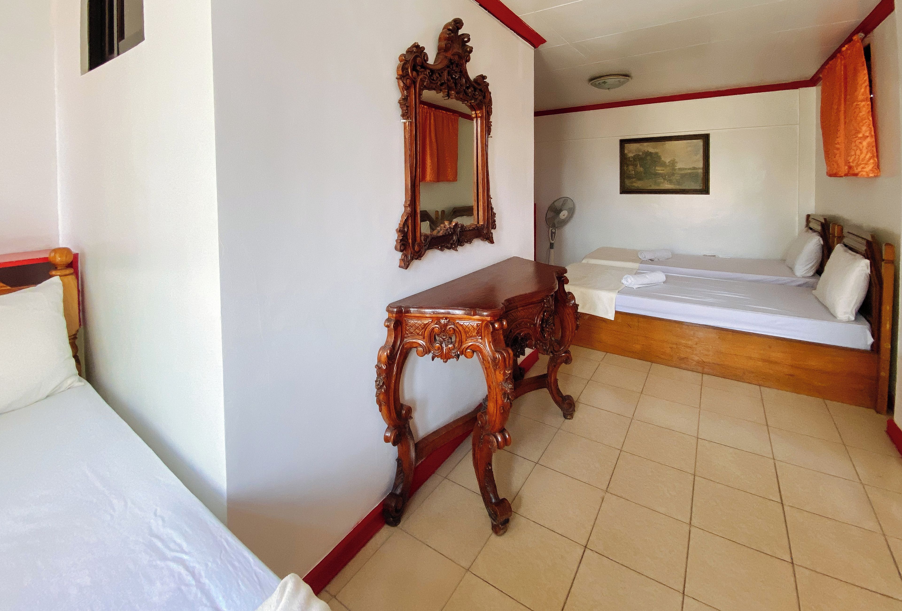 A hotel room with a bed, a wooden nightstand, a mirror, a bedside table, a fan, and a painting on the wall.