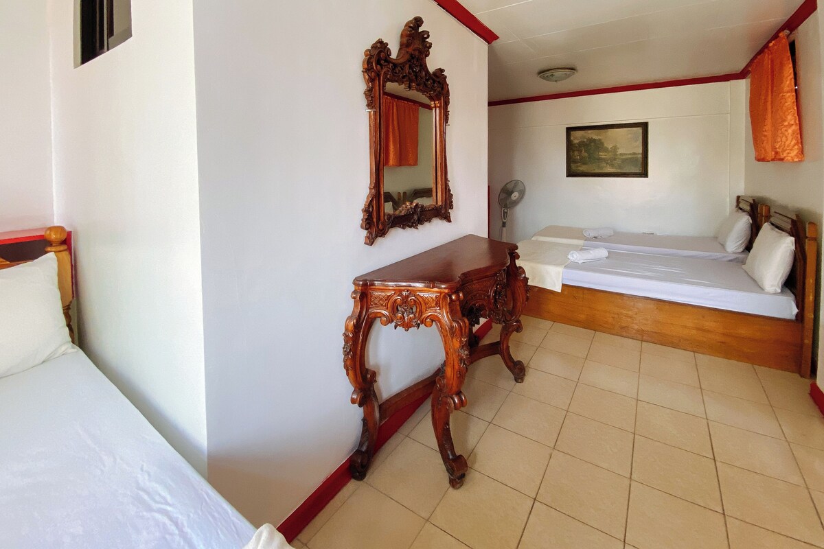 Una habitación de hotel con cama, mesita de noche de madera, espejo, tocador, ventilador y un cuadro en la pared.