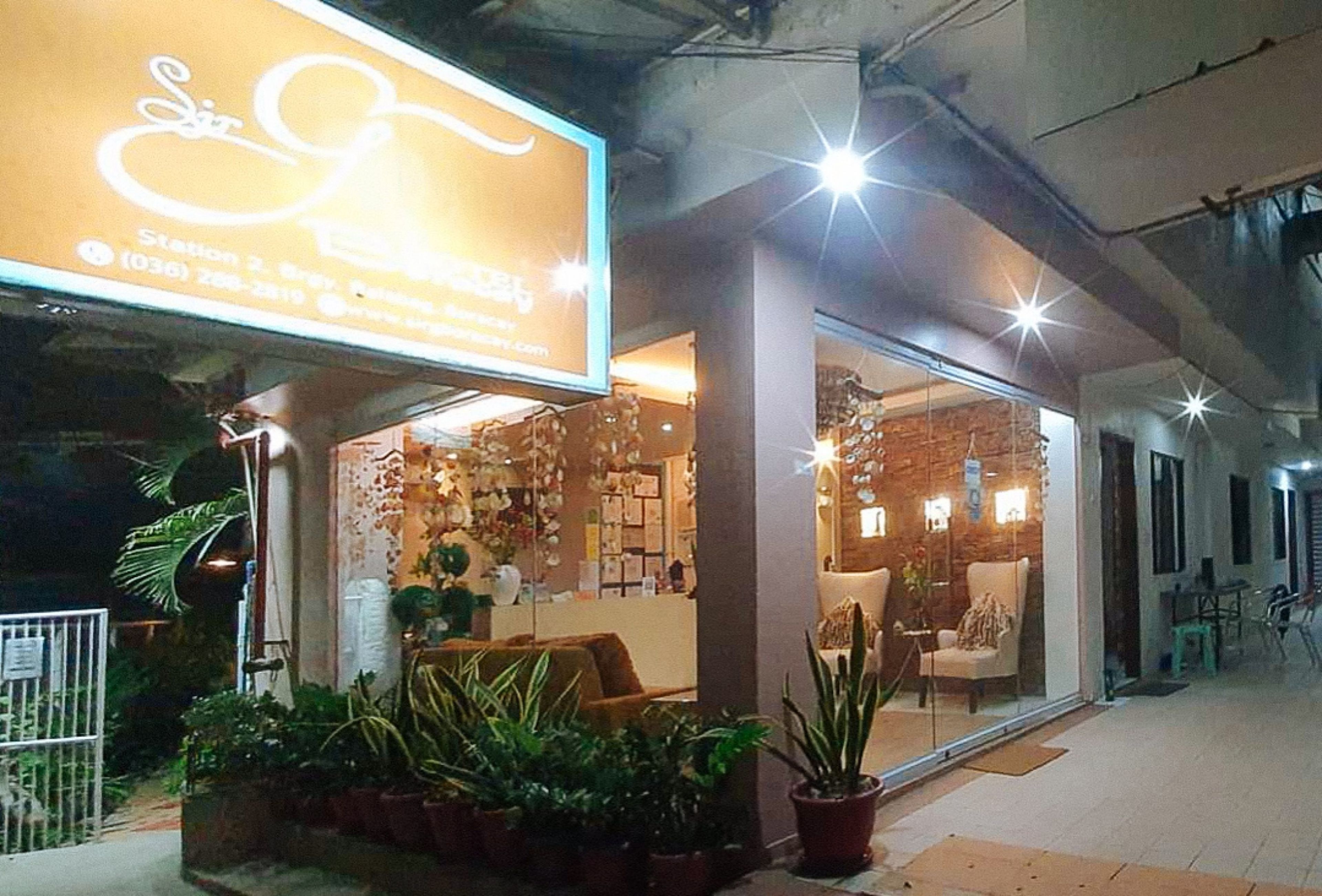 Fachada con un letrero iluminado grande, una zona de recepción con mostrador y un área de estar con sillas y mesas.