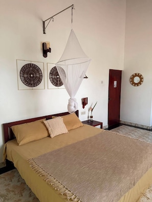 A bedroom with a bed, a wall-mounted mosquito net, two wall art pieces, a wooden door, and a small table with a vase. - Riverbank Bentota (Aluthgama)