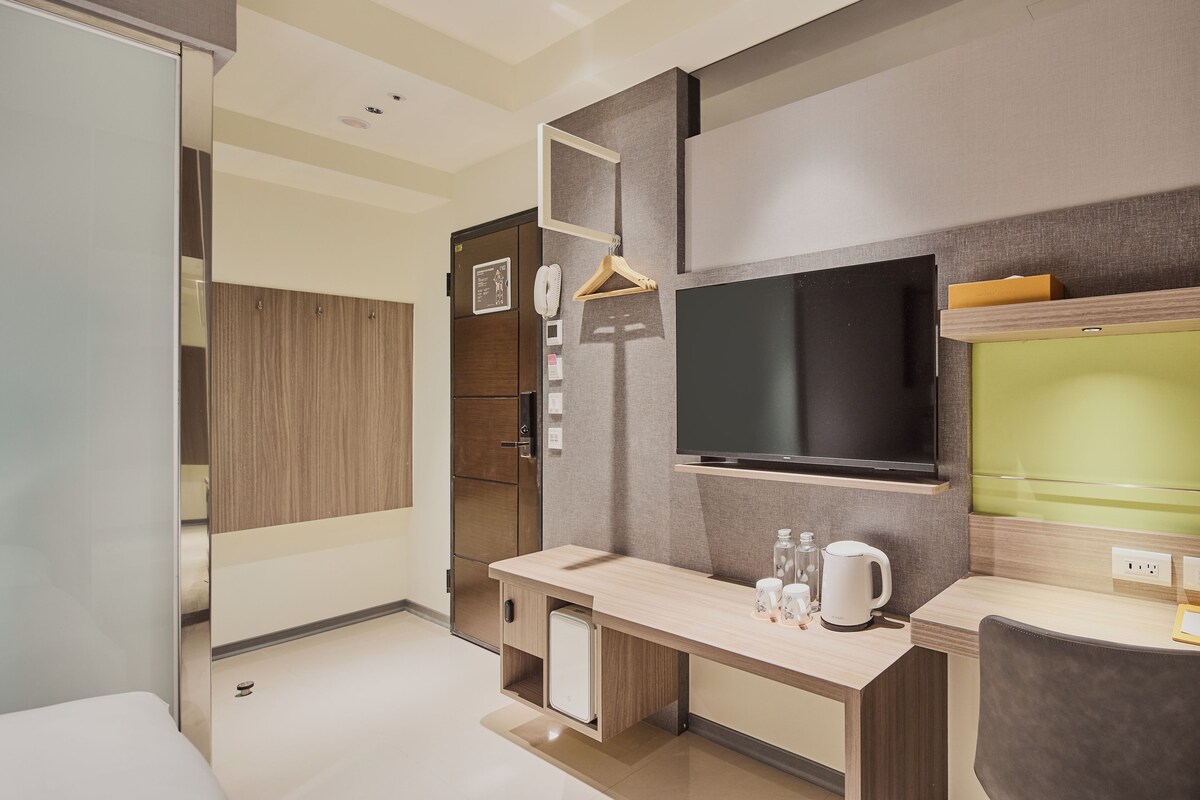 A modern hotel room with a large flat-screen TV, a wooden cabinet, a small seating area, and a bedside table with a kettle and cups.