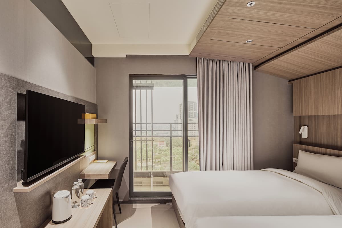 Habitación de hotel moderna con una cama grande, un televisor de pantalla plana en la pared, un escritorio con cafetera y vistas a la ciudad a través de un ventanal amplio.