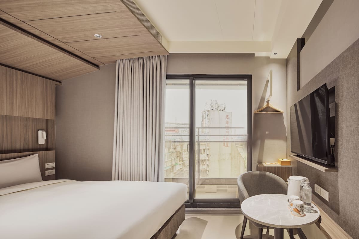 Habitación de hotel moderna con una cama grande, una mesa de comedor, un televisor y un balcón con vistas a la ciudad.